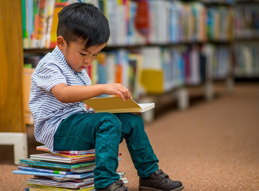 Young child sitting on a stack of books reading in a library aisle